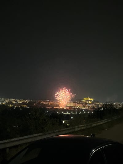 The image shows a nighttime aerial view of a city with fireworks. In the foreground, the dark silhouette of a car is visible, with its roof and part of the windshield illuminated by ambient light. A concrete barrier with graffiti runs along the side of the road in front of the car. Beyond the car, a dark, tree-covered hillside slopes down towards a densely populated urban area. The city is lit by numerous streetlights and building lights, creating a warm glow. In the distance, a prominent hill is topped with a brightly lit structure, possibly a castle or historical monument. A large, vibrant fireworks display erupts in the sky above the city, with bursts of red and yellow light. The sky is dark and clear. The location of La Pedrera, Spain, is not discernible from the visual information in the image.