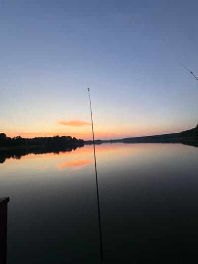 The image depicts a serene sunset scene over a calm body of water, likely a lake, viewed from a dock. The sky transitions from a soft blue at the top to warm hues of orange and pink near the horizon, where a silhouetted treeline marks the distant shore. The water's surface perfectly reflects the sky and the trees, creating a mirror-like effect. A fishing rod stands vertically in the foreground, its tip adorned with a green and yellow bobber. The overall atmosphere is tranquil and peaceful, suggesting a quiet evening by the water. The provided location, Połom, Poland, fits the description of a rural or semi-rural area with natural landscapes like lakes and forests, consistent with the image. No people are visible in the image.