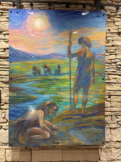 The image is a painting displayed on a stone wall. The painting depicts a prehistoric scene with two human figures and several mammoths in a landscape. In the foreground, a figure is kneeling by the water, seemingly examining something in their hands. In the middle ground, a standing figure holding a spear looks out towards the horizon. Three mammoths are visible in the background, wading through shallow water. The sky is rendered in vibrant colors, with a large sun and swirling brushstrokes suggesting movement. The painting is framed by a dark border and attached to the wall with visible hardware. The stone wall appears to be part of an interior or exterior structure. The location is identified as Unknown, United States of America.
