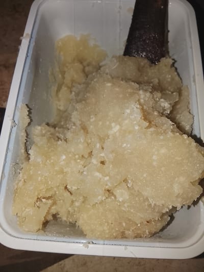 The image shows a close-up, overhead view of a white plastic container filled with mashed or grated food, possibly cassava or yam. The food is a pale yellow color and has a slightly lumpy texture with visible white specks. A dark, rough object, appearing to be a grater or a portion of a tool, is partially submerged in the food on the right side of the container. The container's rim is visible and appears to be slightly wet. The background is blurred, but suggests a countertop surface with brown and beige tones. The scene is set in Rumuokwuta, Nigeria, where such food preparations are common.