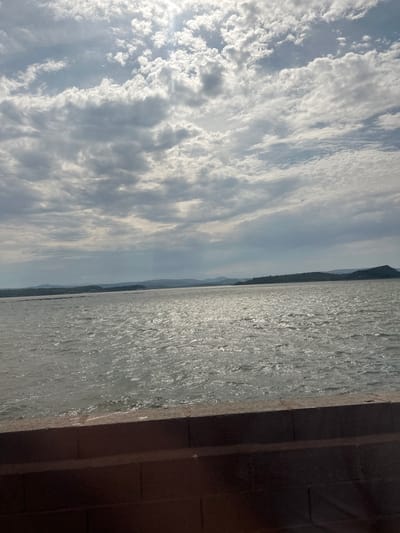 The image depicts a scenic view of a body of water, possibly the Mediterranean Sea, near Port-la-Nouvelle, France. The orientation of the image is vertical, with the sky occupying the left portion and the water and a concrete structure on the right.
The sky is filled with dramatic clouds, some appearing fluffy and white, catching the sunlight, while others are more diffused and grey, suggesting a dynamic weather pattern. The sun is not directly visible, but its presence is indicated by the bright illumination of the clouds. The time of day appears to be late morning or early afternoon, given the quality of light.
The water is choppy, with ripples and glints of sunlight reflecting off its surface, creating a textured, shimmering effect. The vast expanse of water suggests a coastal or open water setting. In the distance, a faint outline of hills or mountains can be seen along the horizon, hinting at a landmass.
On the right side of the image, a dark, textured concrete structure is prominent. It appears to be a wall or part of a pier or embankment, with distinct vertical lines suggesting construction or seams. This structure creates a strong contrast with the natural elements of the sky and water. The overall mood is one of natural beauty and perhaps a sense of stillness despite the textured water. There is no visible text in the image.