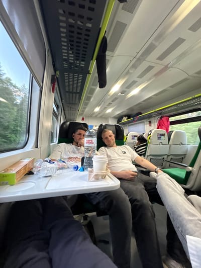 Two men are seated across from each other on a train. One man, wearing a white shirt, is lying back. The other man, also wearing a white shirt, is seated upright. A tray table sits between them with two water bottles, a cup, and a box on it. Other passengers are visible in the background. The view from the train window shows trees. The location is a train traveling through Мажэйкаўскі сельскі Савет, Belarus.