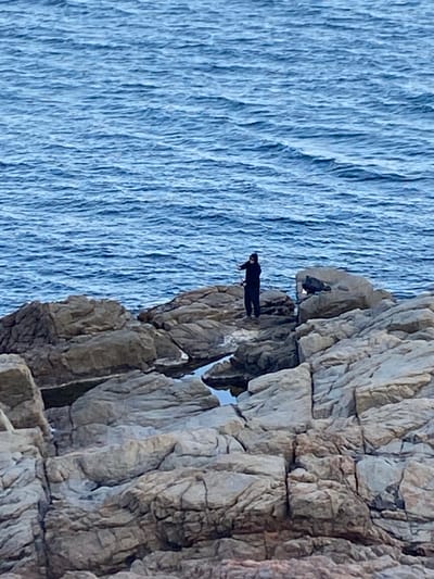 The image shows a person tightrope walking on a rope stretched between rocky cliffs near the sea. The person is dressed in black and appears to be using a balancing pole. The sea is visible on the left. The location is most likely in Palafrugell, Spain, given the prompt's instructions.