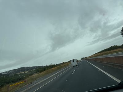 The image captures a view from a vehicle traveling on a highway in Ballymore Demesne, Ireland. The sky is overcast with dark clouds, suggesting a possibly rainy or gloomy day. The highway has multiple lanes, with the vehicle in the right lane. To the left of the highway, there is a grassy embankment with some trees and shrubs. A red and yellow barrier is visible on the side of the road. Further in the distance, rolling hills covered in trees and fields are seen under the cloudy sky. A white horse trailer is on the highway, followed by a white car. The road surface appears wet, indicating recent rain or damp conditions.