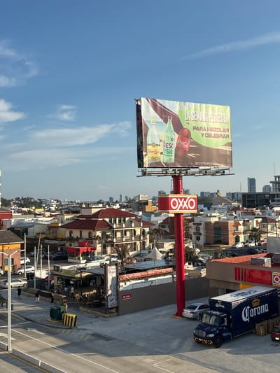 The image is an elevated, urban scene in Boca del Río, Mexico, featuring a variety of elements within the cityscape. The sky is blue with wispy clouds. A large billboard displays an advertisement for Coca-Cola, Fresca, and Topo Chico. Below the billboard, an OXXO convenience store sign is prominently visible. Adjacent to the OXXO is a Corona beer delivery truck and a parked silver sedan.
The scene includes a mix of commercial and residential buildings, some with red roofs. People are visible walking along the sidewalk near a restaurant, "Pulpo Frito," and the surrounding street. A street light and yellow-and-black bollards are also present.