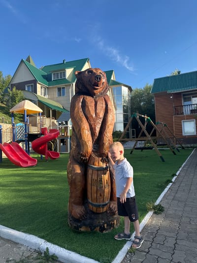 A young boy with blond hair stands to the right of a large wooden carving of a bear holding a barrel. The boy is wearing a light blue polo shirt, black shorts, and gray sandals. He is smiling and has his right hand touching the bear's head. The bear carving is brown and appears to be made of wood. The barrel has the word "МЕД" carved into it, which means "honey" in Russian.
In the background, there is a playground with red slides and a yellow and blue play structure. To the right of the playground, there are two swings. Behind the playground and swings, there are buildings with green roofs. The building on the left appears to be a hotel or guesthouse with beige and brown siding and multiple windows. The building on the right is a wooden structure with a balcony and windows.
The scene is set on a bright, sunny day with a clear blue sky. The ground is covered in green artificial turf, and there is a paved walkway to the right of the grass. The location is identified as Danilovka (Altai), Russia.