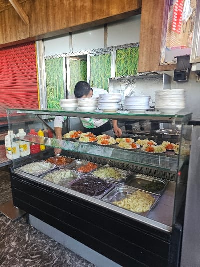 The image displays the interior of a food service establishment located in Boufakrane, Morocco. In the midground, a male individual is positioned behind a counter, leaning forward with his head down, appearing to arrange or prepare food. He is wearing a light-colored shirt. On the counter in front of him are multiple stacks of white ceramic bowls and smaller white plates, each containing individual portions of prepared mixed salads or side dishes.

In the foreground, a rectangular black food display case with a clear glass top presents various cold food items in multiple metal and plastic trays. These items include white grains, orange-colored vegetables, dark leafy greens, a dark solid substance, and light-colored pasta or potato salad. To the left of the display case, on a lower shelf, are three squeeze bottles (two yellow, one red) and two larger clear plastic bottles.

The background features a wall section adorned with green wallpaper depicting a dense grass or reed pattern. A silver-framed doorway is visible within this wall. To the far left, a red roll-down security shutter is partially visible, indicating an exterior opening. Wooden beams are present overhead. The floor exhibits a dark, marbled pattern.