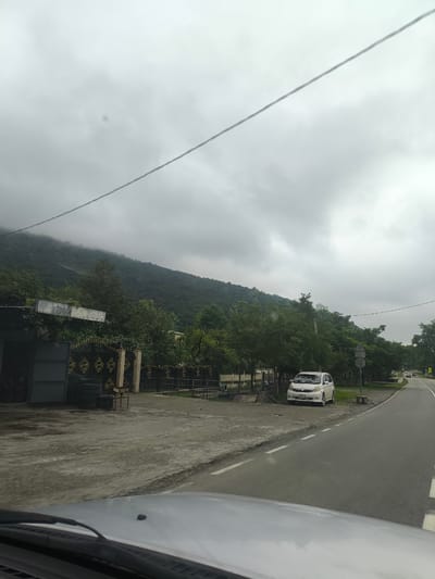 The image depicts a roadside scene in Gachedili, Georgia, under an overcast sky. In the foreground, the hood and windshield wipers of a silver vehicle are visible. A paved road with dashed white lines runs diagonally through the frame from right to left. On the left side of the road, there is a gray, open-sided structure with a metal roof, possibly a workshop or storage area, where stacks of tires are present. Adjacent to this structure is an ornate black metal gate with gold detailing, leading to an unseen property. Behind the gate, various trees and foliage are visible. Further along the road, a white minivan is parked on the side. In the background, a densely forested mountain slopes upwards, its peak partially obscured by clouds. The overall environment appears to be rural or semi-rural, with a road infrastructure and natural landscape.