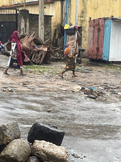 In the city of Unknown, Unknown, two women are walking on a dirt road that has puddles of water, suggesting it might be raining or has recently rained. The ground is a mix of dirt, gravel, and debris. In the background, there are buildings and a large metal shipping container.
One woman, on the left, is wearing a red headscarf and a patterned dress. She is walking towards the left side of the frame. The second woman, on the right, is carrying a black basin on her head filled with items, including a yellow object. She is also wearing a patterned headscarf and a patterned skirt, and has a baby on her back. She is walking towards the right side of the frame.
The scene is cluttered with debris, including large pieces of wood and scattered trash. The overall impression is of a working-class or impoverished urban environment.