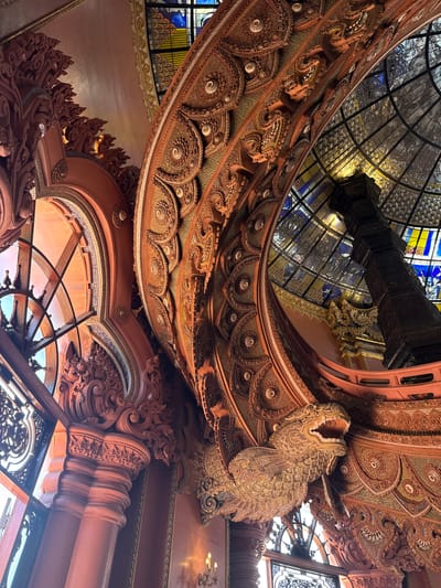 This low-angle shot shows the intricately decorated interior of the Erawan Museum in Samrong Nuea Subdistrict Municipality, Thailand. Elaborate carved details adorn the walls, ceiling, and supporting structures. The carvings are primarily reddish-brown with ornate patterns, including circular motifs and depictions of mythical creatures. Above, a stained glass ceiling with a complex geometric pattern allows light to filter into the space. A dark, boxy structure hangs from the center of the ceiling. The architecture displays a mix of traditional Thai and Western influences, evident in the decorative elements and structural design. The overall atmosphere is one of grandeur and reverence.