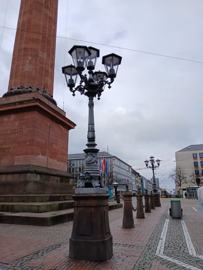 The image depicts the Luisenplatz in Darmstadt, Germany, under an overcast sky. Dominating the foreground is the Luisen Monument, a tall red sandstone pillar with an ornate lamp post attached to the top. The base of the monument is adorned with inscriptions and decorative elements.

Adjacent to the monument is a brick-paved pedestrian area lined with bollards connected by chains. In the background are buildings, including a modern glass-fronted structure with flags. A tram is visible further down the street. There are no people visible in the immediate area.