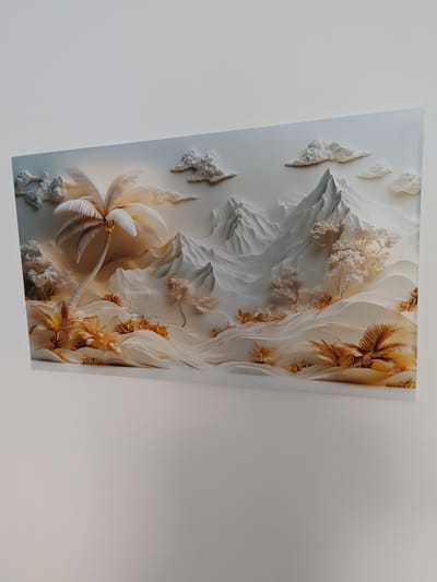 This is a horizontal, medium shot of a framed piece of art depicting a monochromatic, stylized tropical mountain landscape. The artwork is mounted on a white wall. The scene is rendered in shades of white, cream, and pale gold, with a textured, almost sculptural appearance.
The foreground features rolling sand dunes and sparse, stylized vegetation, including palm fronds and small, feathery trees. A prominent, tall palm tree with a textured trunk and bright white fronds stands to the left of the composition. In the midground, abstract, pointed mountain peaks rise dramatically, rendered with sharp angles and smooth surfaces. Wispy, cloud-like formations are scattered across the sky.
The overall impression is serene and ethereal, with a soft, warm light illuminating the scene, suggesting either sunrise or sunset. The use of monochromatic tones and the textured, relief-like style give the artwork a unique, almost ethereal quality. There is no visible text in the image. The setting appears to be an indoor display of art, with the artwork itself being the sole subject.