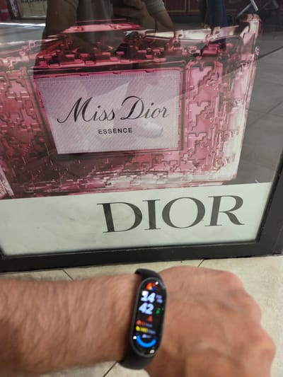 The image shows a close-up of a person's left wrist wearing a black fitness tracker. The tracker displays a digital watch face with time 14:42 and various icons. In the background, a large advertisement for "Miss Dior Essence" and "DIOR" is visible, reflected in a glass surface. The advertisement features a pink, textured bottle and the brand name in prominent lettering. The setting appears to be an urban environment, potentially a storefront or display window. The flooring is tiled. The presence of the advertisement suggests this image may have been taken in a commercial area. Without additional information, the specific location within Collado Villalba, Spain, cannot be determined.