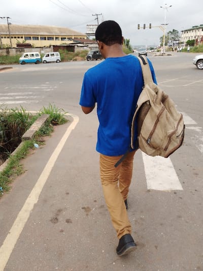 The image captures a street scene in Irete, Nigeria, under an overcast sky. A male individual, seen from behind, walks forward on a paved surface. He is dressed in a blue short-sleeved t-shirt, tan trousers, and dark shoes. A tan fabric single-strap backpack with brown leatherette accents is worn over his right shoulder. To his left, a yellow line marks the edge of the asphalt, adjacent to an open concrete drainage ditch largely filled with green vegetation. Further to the right on the pavement, a partial white rectangular marking, consistent with a pedestrian crossing, is visible. The asphalt exhibits several dark stains.

In the mid-ground, across the road, two small white vans are parked, one featuring blue detailing. Behind them stands a large, light-colored industrial or warehouse-type building with a corrugated roof. Overhead, utility poles and power lines crisscross the sky. Further along the road to the right, a multi-lane intersection is apparent, equipped with traffic lights suspended from gantries and a prominent streetlight fixture. Vehicles, including a dark-colored car and a silver SUV, are present on the roadways. Distant buildings and vegetation extend towards the horizon, indicating an urban setting.