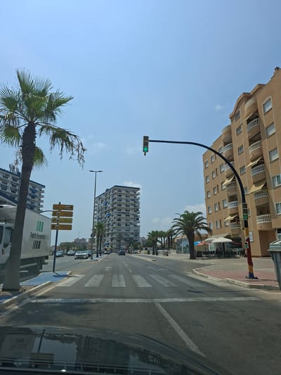 The image shows a street scene in La Manga Del Mar Menor, Spain, on a clear day with a blue sky. A wide, paved road with multiple lanes and a pedestrian crossing is visible. Traffic lights are present, with a green light illuminated on the main signal. Palm trees line the street, some of which are mature and tall. Modern, multi-story apartment buildings with balconies are prominent on both sides of the road. On the left side, a white truck with green lettering that reads "Petit Forestier" is parked. Yellow directional signs indicating hotels such as "Hotel Benidorm La Manga," "Hotel Doblemar," "Hotel Bahia La Manga" and "Aparthotel Londres" are also visible. Cars are driving on the road. On the right side of the road, there are outdoor seating areas for a cafe or restaurant with umbrellas providing shade. The overall environment suggests a coastal resort town.