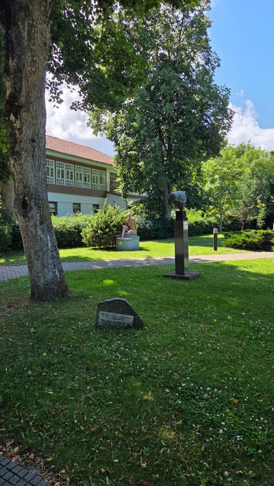 A statue of a nude woman sitting on a circular base is located in a park in Palanga, Lithuania. The statue is made of bronze and is approximately life-size. The woman is depicted with her legs crossed and her head tilted back. Behind the statue is a building with a red tile roof and a row of white windows. In front of the statue, on the grass, is a stone with an inscription in Lithuanian. There is also a sculpture of a bird's head on a black pedestal. The park is well-maintained, with lush green grass and trees. The sky is blue with some clouds.