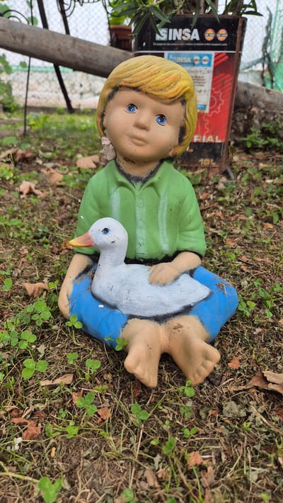 A garden gnome, depicting a young child with blonde hair, blue eyes, and a green shirt, sits on the ground holding a white duck. The gnome is positioned outdoors, surrounded by dry grass, fallen leaves, and small green plants. In the background, a metal post and a black container with red and white lettering are visible. The scene appears to be located in an outdoor setting, possibly a garden or yard. The precise location in Doirentsi, Bulgaria, is not discernible from the image alone.