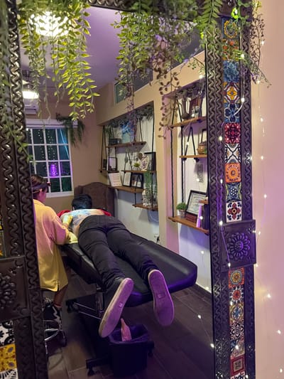 A reflected image shows a person in black pants and purple sneakers reclining in a black salon chair. The reflection shows a woman receiving a tattoo, visible in color on her back, in a room with tiled floors and shelves displaying various items. A window with a neon tattoo sign is also visible. The reflection is in a mirror framed with decorative tiles and foliage, including fairy lights. The image was taken in Ciudad Madero, Mexico.