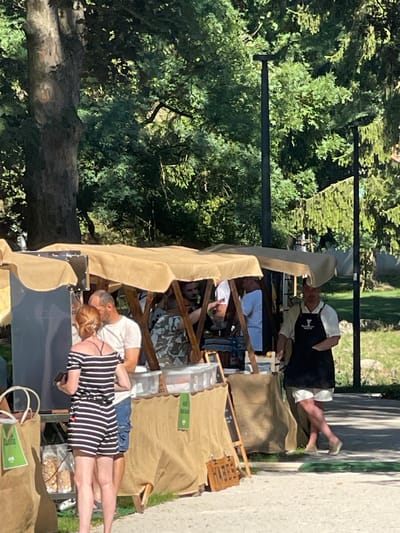 The image depicts an outdoor market scene in Stupava, Slovakia, characterized by several stalls with beige awnings. In the foreground, a woman wearing a black and white striped off-the-shoulder romper stands with her back to the camera, holding a black object in her left hand. To her right, a man in a white t-shirt and blue shorts stands beside her. Behind them, several other individuals are visible within the market stalls, some interacting with goods or each other. One man, wearing a white shirt and a black apron, is seen standing to the right of the stalls. The market is set against a backdrop of lush green trees and foliage, with lampposts visible in the background. The ground appears to be a paved pathway.