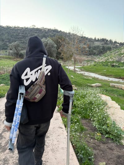 An individual, viewed from behind, walks on a light-colored concrete or stone path in the Emek Izrael Regional Council, Israel. The person wears a black hooded sweatshirt with a prominent white "STUSSY" logo on the back, dark trousers, and white wristbands. A colorful, striped fanny pack or sling bag is worn across the back. The individual uses a silver forearm crutch in their right hand for support, and a second folded silver forearm crutch, featuring a blue and white patterned accessory, is secured to their left side. To the right of the path, green grass is dotted with numerous small white flowers, and a narrow stream or water channel flows parallel. The background features green, undulating hills sparsely covered with mixed evergreen and deciduous trees, with several light-colored buildings visible on a distant hilltop. The sky is pale blue, indicating daylight.