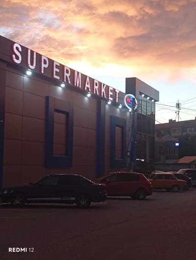 The media file shows a building with a large sign that reads "SUPERMARKET." The building appears to be a store, with large windows and a modern facade. There are several cars parked in front of the building, including a red car and a black car. The sky in the background is a dramatic orange and pink, suggesting it is either sunrise or sunset. The lighting indicates it is dusk. The overall scene is an urban setting, possibly a street in a town or city. The camera is angled upwards, making the building and the sky prominent. There is text on the right side of the image that reads "REDMI 12," likely indicating the device used to capture the image. The weather appears to be clear with dramatic clouds.