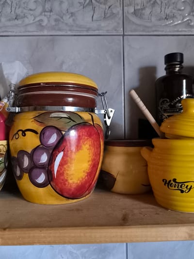 This is a close-up, eye-level, indoor shot of kitchen items on a wooden shelf. The background is a grey tiled wall. On the left is a large, yellow ceramic canister decorated with painted grapes and a peach. It has a brown lid with a metal clasp. To the right of the canister are two smaller yellow ceramic containers, one with a lid, and a dark bottle with a label that reads "RĪGAS MELNĀIS." A light wooden stick leans against the dark bottle. The scene is lit from the front, casting shadows. The image appears to be located in a kitchen in Žiaukos, Lithuania.