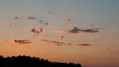 The media file is a photograph of a sunset over a silhouetted treeline. The sky transitions from a pale blue at the top to a warm orange near the horizon. Scattered clouds are illuminated by the setting sun, appearing in shades of pink, orange, and grey. The foreground features a dark, undulating silhouette of trees, indicating a natural landscape. The time of day is clearly sunset, and the atmosphere appears calm and serene. No people, objects, or text are visible in the image. The location context is Strengberg, Austria.