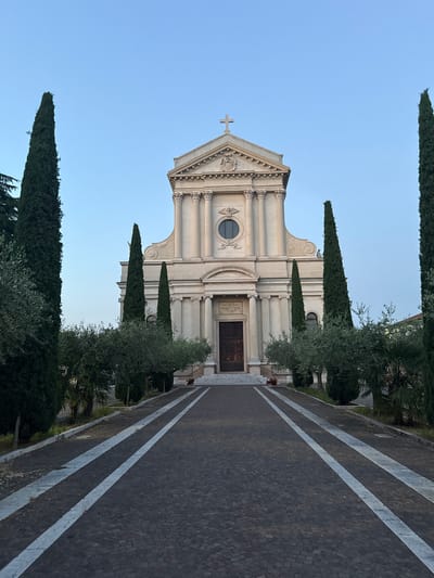 The image shows the Church of Saints Peter and Paul in Villafranca di Verona, Italy. The facade is pale, with a cross at the top. The building has classical architectural features, including columns. The entrance is a large, dark door with a plaque above it. Flanking the church are tall, narrow cypress trees. A paved walkway with white lines leads towards the church. The sky is clear and blue.