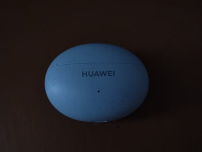 The image shows a closed Huawei earbuds case on a brown surface. The case is round, light blue, and speckled with darker spots. The word "HUAWEI" is printed in dark letters on the top of the case. A small pinhole is visible below the logo. The environment is dark, with the case being the primary lit subject. The image was taken in Дудзіцкі сельскі Савет, Belarus.
