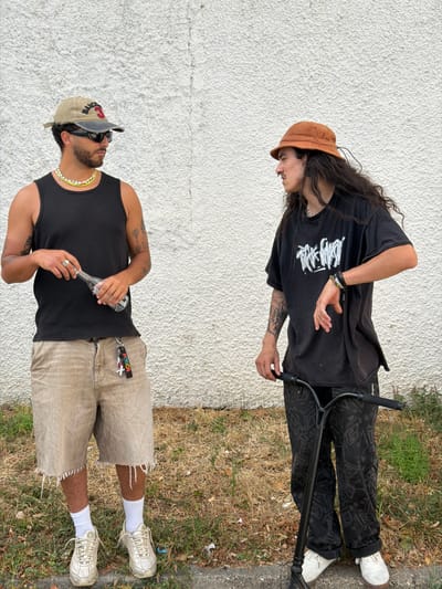 The media file displays two individuals outdoors, against a textured white wall and a grassy embankment. The upper individual, oriented towards the viewer's right, wears a black t-shirt with a white graphic design, black pants with a patterned print, and white shoes. They have long, dark hair and are wearing a brown corduroy bucket hat. Their stance suggests they are interacting with a black scooter. The lower individual, facing the viewer's left, is dressed in a black tank top, khaki shorts with frayed hems, and white sneakers with white socks. They are wearing sunglasses, a beaded necklace, and a ring on their left hand. This person is holding a clear glass bottle. A keychain with the text "POSCA" is attached to their shorts. The lighting suggests it is daytime, and the dry grass indicates warm weather. The overall scene appears to be casual and urban, possibly a street or sidewalk area in Caluire-et-Cuire, France.