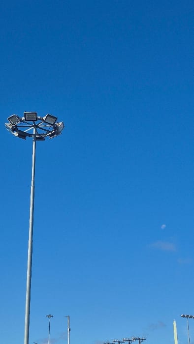 A daytime view captures a vast, cloudless blue sky over Hualpén, Chile. A prominent tall metal pole, topped with a circular array of numerous rectangular spotlights, extends diagonally from the lower-left towards the center of the frame. In the upper-middle section of the sky, a faint, pale moon is visible. To the right, several similar light poles with their own arrays of spotlights are visible in the distance, suggesting a large outdoor venue such as a sports stadium or arena. The clear, bright blue sky indicates fair weather. A subtle, reddish-brown smudge or discoloration can be seen on the right side of the sky, possibly a photographic artifact or a distant atmospheric feature. There is also a very faint, light-colored horizontal streak near the top right edge. No people or text are visible in the image. The time of day appears to be either early morning or late afternoon, when the moon is still perceptible against a bright sky.