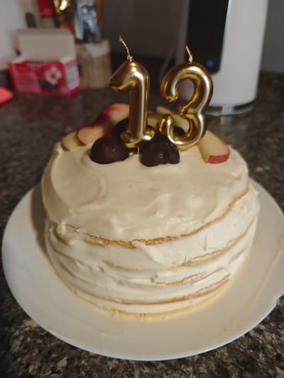 A small, layered cake, frosted with white icing, sits on a white plate on a dark, speckled countertop. Two gold candles, shaped like the numbers 1 and 3, are stuck into the top of the cake. The cake is decorated with slices of what appear to be apple or peach and dark cherries. In the background, a white kitchen appliance, possibly a blender or coffee maker, is visible. To the left of the appliance, a partially visible pink box with floral designs is on the counter. The image is well-lit, with natural light illuminating the cake. The location is not specified in the image.