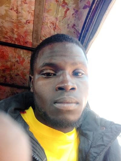 The image presents a headshot of a dark-skinned male with short dark hair and a short beard, facing forward with a neutral expression. He is wearing a bright yellow t-shirt beneath a dark, zippered jacket, with a portion of a blurred digit visible in the lower-left foreground. The background on the left features patterned fabric, exhibiting floral motifs in shades of orange, red, brown, and purple, bisected by a vertical brown structural strip. The background on the right is an overexposed white area, indicative of bright outdoor lighting. This scene is situated in Rijiyar Lemo, Nigeria.