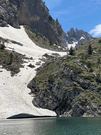 This forensic description details an outdoor scene in the city of Shalë, Albania, featuring a mountain lake, snow-covered slopes, and prominent rock formations under a clear sky.

The lower portion of the image is dominated by a deep teal-colored mountain lake, its surface rippled by a gentle breeze. Along the left edge of the lake, a substantial snowbank extends into the water, forming a thick, overhanging edge. Above the lake, on the left, a steep mountain slope is heavily covered with white snow, which exhibits textured patterns and partially exposed patches of dark earth and sparse vegetation. Interspersed throughout the snow-free areas of this slope and the adjacent rocky terrain are scattered small evergreen trees.

On the right side of the lake, a rugged, rocky slope ascends steeply, covered with green grasses, low shrubs, and numerous gray rock outcrops and visible bedrock strata. Several small evergreen trees are also present on this vegetated slope.

In the mid-ground and background, the landscape transitions to imposing rock formations. A massive, sheer gray rock face rises vertically on the left, displaying geological layering and crevices. Towards the upper right, a series of jagged, pointed mountain peaks are visible against the blue sky, with patches of snow remaining in their depressions. One of these peaks prominently features a distinct, natural arch-like opening. The sky above is a bright, clear blue, with minimal, wispy white cloud cover on the right side. No people, animals, or artificial structures are discernible within the frame.