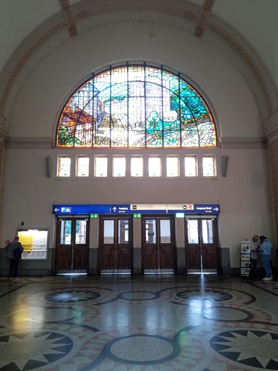 The image shows the interior of the Eisenach train station. Dominating the upper half of the frame is a large, arched stained-glass window depicting a stylized landscape or cityscape with buildings and greenery. Below the stained-glass window, a series of smaller rectangular windows are visible. The central part of the image features a wall with several wooden doors. Above the doors are blue signs with directional arrows and text in German. One sign indicates "Ausgang Stadt" (Exit City) with an arrow pointing to the right. To the left of the doors, there is a large bulletin board or display with information. The floor is decorated with a geometric patterned mosaic. In the foreground, the light from the stained-glass window reflects on the polished floor, creating colorful patterns. On the left, a person wearing a grey jacket and dark pants stands facing the bulletin board. On the right side, near a display rack with pamphlets, two individuals are engaged in conversation. One person is wearing a light blue jacket and dark pants, and the other is wearing a grey shirt and dark pants. The overall environment appears to be a public indoor space, likely a waiting area or concourse within the train station.