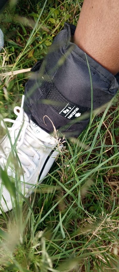 A close-up, downward-angled view depicts a person's lower leg and foot, positioned within a dense, green grassy environment. A black fabric ankle weight, labeled with "SUZUKI" (including a stylized 'S' logo) and "1 Kg" in white text, is secured around the wearer's light brown-skinned ankle. Beneath the ankle weight, a white athletic shoe or sneaker with white laces is visible, featuring a black stripe along its side. Faint text, possibly "ok. 17", is partially visible on the shoe's upper material. The foreground and background are filled with various blades of green and some dried grass, suggesting an outdoor, unmanicured setting.
