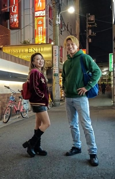 Two smiling young adults, a female and a male, stand side-by-side on an asphalt street at night in Gifu, Japan, posing for a photograph. The female, on the left, has long brown hair with a shaved side and wears a maroon velvet bomber jacket with gold trim, a black miniskirt, black mid-calf platform boots, and a red backpack. The male, on the right, has blonde hair and wears a dark green hooded sweatshirt, light blue jeans, and black sneakers, with a blue tote bag slung over his left shoulder and his left hand on his hip.

Behind the individuals, several multi-story buildings line the street, featuring prominent illuminated signage. A large golden sign for "HOTEL ENIU" is visible on a building to the left, with smaller Japanese text underneath. Numerous vertical neon signs in red and orange with Japanese characters also illuminate the scene. A utility pole with attached wires and a functioning streetlamp stands between the subjects and the buildings. A pink bicycle with a front basket and kickstand is parked to the left, behind the female. A blurry figure is discernable in the distant background, further down the street.