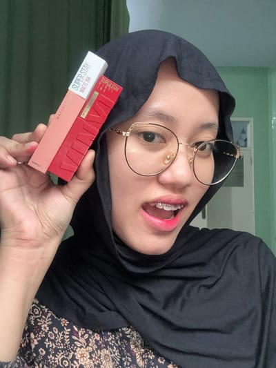 An individual is depicted indoors in Patrang, Indonesia, holding two boxed Maybelline cosmetic products. The person, a young female, is wearing a black hijab, round gold-rimmed eyeglasses, and a dark floral-patterned garment. She holds a white and peach-colored "MAYBELLINE SUPER STAY MATTE INK" box above a red "MAYBELLINE VINYL INK" box in her left hand, which is positioned in front of her right shoulder. Her mouth is open, revealing clear orthodontic braces on her upper and lower teeth, and her lips are covered in bright pink lipstick. Her eyes are directed towards the camera. The background consists of a dark green fabric on the left and a light green wall on the right, with a faint architectural detail visible on the far right.
