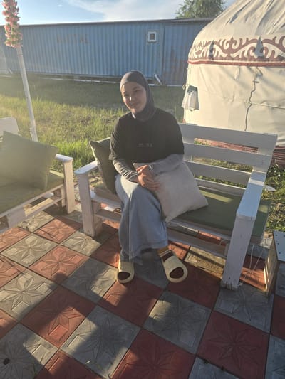A woman wearing a hijab and a black long-sleeved shirt sits on a white wooden bench outdoors. She is holding a beige pillow and wearing light blue jeans and yellow and white striped slippers. The bench is made of wooden slats and has green cushions. Behind the bench is a large, cream-colored yurt with a decorative pattern along the top edge. To the left of the yurt is a blue shipping container. The ground in front of the bench is paved with red and gray square tiles, each featuring a star pattern. Lush green grass and some trees are visible in the background. The sun is low in the sky, casting long shadows and illuminating the scene with warm, golden light, suggesting it is late afternoon or early evening. The overall atmosphere appears relaxed and peaceful. There is no visible text in the image.