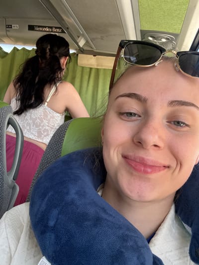 A front-facing, close-up selfie captures a young woman with fair skin, freckles, and light green eyes, wearing sunglasses on her head and a blue neck pillow. She is smiling and appears to be on a bus, with glimpses of another person with dark hair in a white lace top and pink pants visible in the background. The bus interior features green curtains and a "Mercedes-Benz" logo on the ceiling. The setting is likely within Albufeira, Portugal, given the context of a bus journey in the Algarve region.