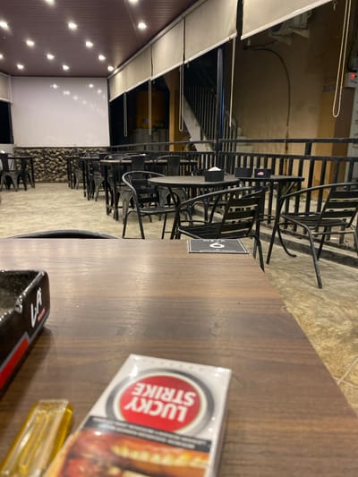 The image depicts a table at an outdoor seating area, likely in Keudeublang, Indonesia. A pack of "Lucky Strike" cigarettes and a lighter are on the table, along with a black tray. In the background, there are numerous black chairs and tables arranged along a walkway with a textured stone wall. The outdoor area is illuminated by several small lights along the roof and walls.