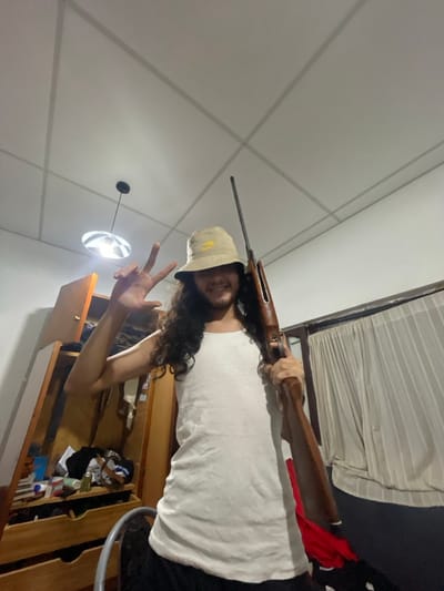 An individual, appearing male with long dark curly hair and a beard, is depicted indoors. He wears a light-colored bucket hat with a yellow logo and a white ribbed tank top. He is smiling and looking towards the camera. His left hand is raised, forming a "V" sign, while his right hand holds a long gun, consistent with a break-barrel air rifle or a small-caliber rifle, featuring a wooden stock. The rifle is held in a near-vertical position. The background shows a room with light-colored walls and a white ceiling with grid lines. To the left, a pendant light fixture hangs, and a wooden wardrobe or dresser with an open door reveals internal shelves containing various items and a reflective surface. To the right, part of a bed frame or bunk bed is visible behind a light-colored curtain, with red and dark-patterned fabrics on the bed. The specific location within the city of Municipio de San Salvador de Jujuy, Argentina, cannot be determined from the visual information provided.