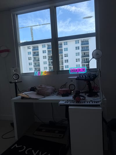 A workspace sits near a window with a view of a tall building and blue sky. A condenser microphone, pop filter, DJ controller, headphones, keyboard, and studio monitors are visible on the desk. Neon signs spelling "STUDIO" and "MUSIC" adorn the space. A black rug with a white graphic and a cassette tape design are positioned next to the desk.