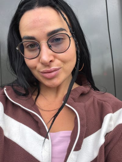 A person with black hair and glasses is wearing a brown and white zip-up jacket over a light purple top. The person has light skin, and the jacket is partially unzipped. The background is a grey surface. No specific location in Sust'e-Konets, Russia, can be determined from the provided image.