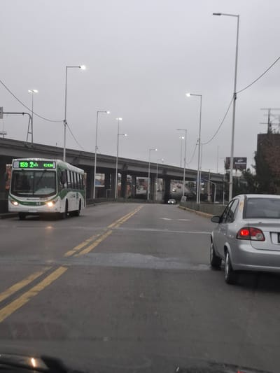 This image captures a street scene in Dock Sud, Argentina, under an overcast sky, with artificial lights illuminated. The perspective is from inside a vehicle, with a slight reflection visible on the lower dashboard.
In the left midground, a white and green public bus, identified as line "159 2 COI" with "Metalpar" branding and the number "44," faces the viewer at a slight angle. Its headlights are illuminated, and a disabled access symbol is visible on its front display.
On the right midground, a silver four-door sedan, appearing to be a Chevrolet Corsa/Classic model, is positioned with its rear facing the viewer, its red taillights illuminated.
The road surface is asphalt, marked by double solid yellow lines. An elevated concrete overpass structure with multiple support pillars spans the background horizontally. Numerous tall, grey street light poles with illuminated LED fixtures line the road, their bright lights creating distinct points against the dim, uniformly grey sky. Additional vehicles are visible on other road levels in the distant background, beneath and beyond the overpass. To the far right, a dark building with antennas and some sparse foliage is partially visible, along with a large billboard above the overpass. The general lighting suggests either dusk, dawn, or a heavily overcast day requiring artificial illumination.
