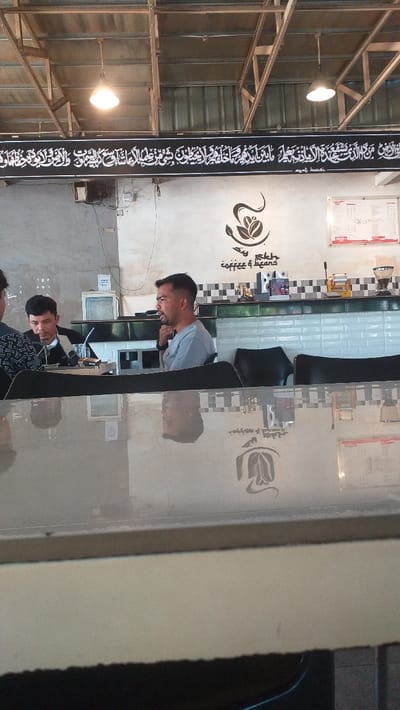 The image depicts an indoor scene within a cafe or coffee shop located in Meuriya, Indonesia. The foreground features a reflective tabletop, mirroring elements of the ceiling and objects beyond it. Three adult males are visible. In the mid-ground, a man with short dark hair and a beard, wearing a light grey t-shirt, is seated at a table, facing left and looking forward. To his left and slightly behind him, another man, wearing a dark jacket over a white shirt, is seated, looking towards the man in the grey t-shirt, with an open laptop visible on the table in front of him. A third man, partially obscured on the far left, wears a patterned shirt and looks downwards.
The background reveals a service counter with a black and white checkered tile backsplash above white subway tiles. Various coffee-making equipment, a speaker, and other items are arranged on the counter. A "coffee & beans" logo, featuring a stylized coffee bean and leaf, is painted on the wall above the counter. Further above, a long black banner with white Arabic script spans the wall. The ceiling features an exposed industrial design with corrugated metal sheeting, metal beams, and wooden supports. Two pendant lights with illuminated bulbs hang from the ceiling structure. A white menu or notice board is affixed to the right wall. Black chairs are visible behind the seated individuals.