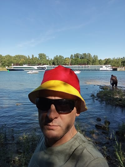 A man wearing a German flag-themed bucket hat and sunglasses takes a selfie by the Rhine River in Biesheim, France. The river, a deep blue with gentle ripples, occupies the middle ground, with a large white passenger ship and a smaller white boat visible on its surface. The background features a lush green tree line under a clear blue sky with a few wispy clouds. On the right bank of the river, a person in dark shorts and a brown shirt is partially visible, facing away from the camera and appearing to bend over. The man in the foreground is wearing a muted green t-shirt and his face is partially obscured by his hat and sunglasses. The scene captures a sunny day at the riverfront.
