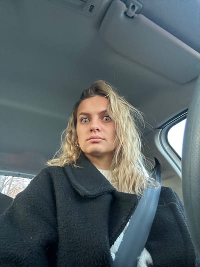 A woman with blonde, wavy hair and striking green eyes sits in a car, looking directly at the camera. She's wearing a white top and a thick, black wool coat. The car's interior is visible, including a grey sun visor and the dark grey seatbelt draped across her chest. The lighting is bright, suggesting it's daytime. The angle is slightly tilted.