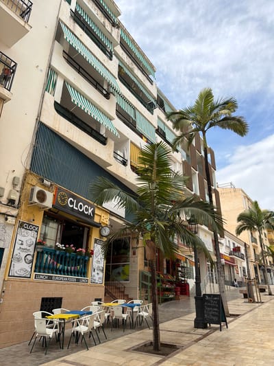 The image shows a street scene in Arroyo De La Miel-Benalmadena Costa, Spain. In the foreground is a cafe named "CLOCK," with an awning, menu board, and outdoor seating consisting of white chairs with colored (yellow and blue) fabrics. The cafe's exterior is decorated with flowers, and a clock is visible above the entrance. Adjacent to the cafe are tall buildings, one with green and white striped awnings. Palm trees are positioned along the street and partially obscure the view of the buildings. A black board with Russian menus is partially visible and near the cafe. The environment is typical for a Mediterranean coastal city, with a mix of commercial and residential buildings and the presence of palm trees. The sky is partially cloudy.