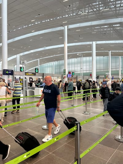 The image depicts the interior of an airport terminal in Granadilla, Spain. The scene is a check-in area where numerous passengers are waiting in line. The passengers are standing behind a yellow and green rope barrier. A man is pushing a luggage cart, which is filled with a black suitcase. The environment is a bright, well-lit terminal with white walls and a modern ceiling design. The floor is tiled with grey tiles. There are multiple check-in counters with information signs visible on the walls.  The people appear to be waiting for their flights. There are more people and luggage in the background. The scene is in the Tenerife South Airport (TFS), located near the town of Granadilla, Spain. The latitude and longitude coordinates, 28.047 and -16.58, respectively, confirm that the scene is indeed at the Tenerife South Airport.